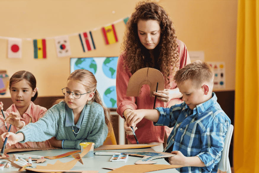 Lehrerin bastelt mit Kindern im Klassenzimmer – frühe Finanzbildung und Familienbudget mit Kindern spielerisch erklären.