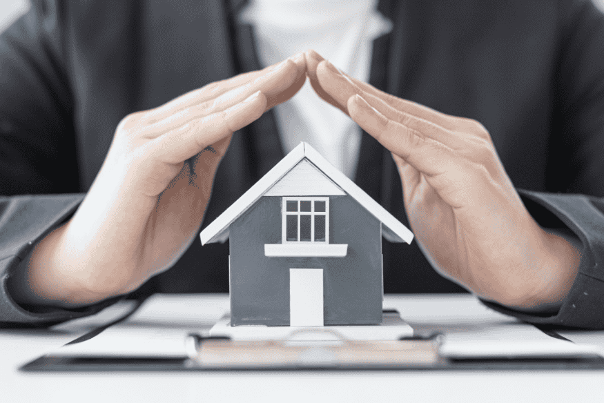 Person schützt ein Modellhaus mit den Händen – Konzeptbild für Hausratversicherung und Vermögensschutz.