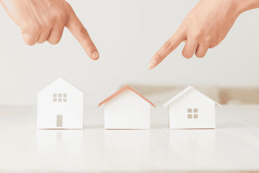 Finger zeigen auf verschiedene Modellhäuser – Auswahl der passenden Immobilie als Geldanlage im Vergleich zu anderen Objekten.
