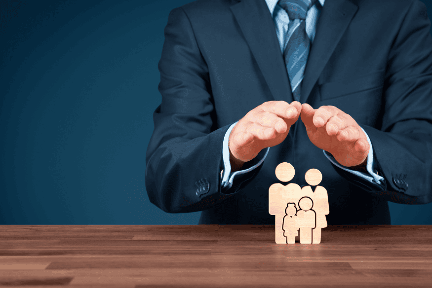 Geschäftsmann hält seine Hände schützend über Holzfiguren einer Familie, symbolisch für Pflege- und Risikoabsicherung.