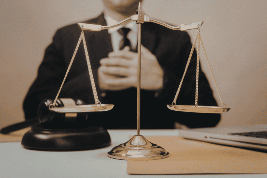 Justitia-Waage auf einem Schreibtisch mit Anwalt im Hintergrund.