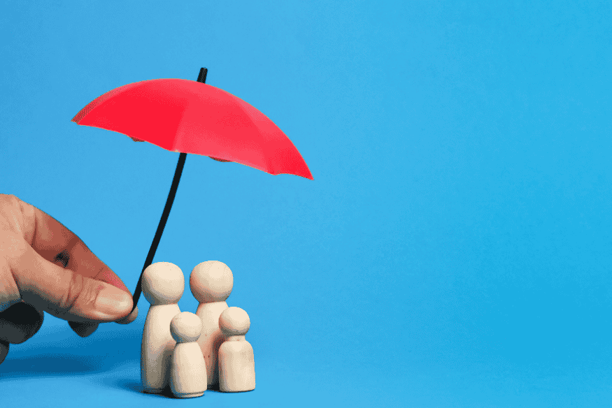 Hand hält einen roten Schutzschirm über einer Holzfigur-Familie als Symbol für finanzielle Absicherung.