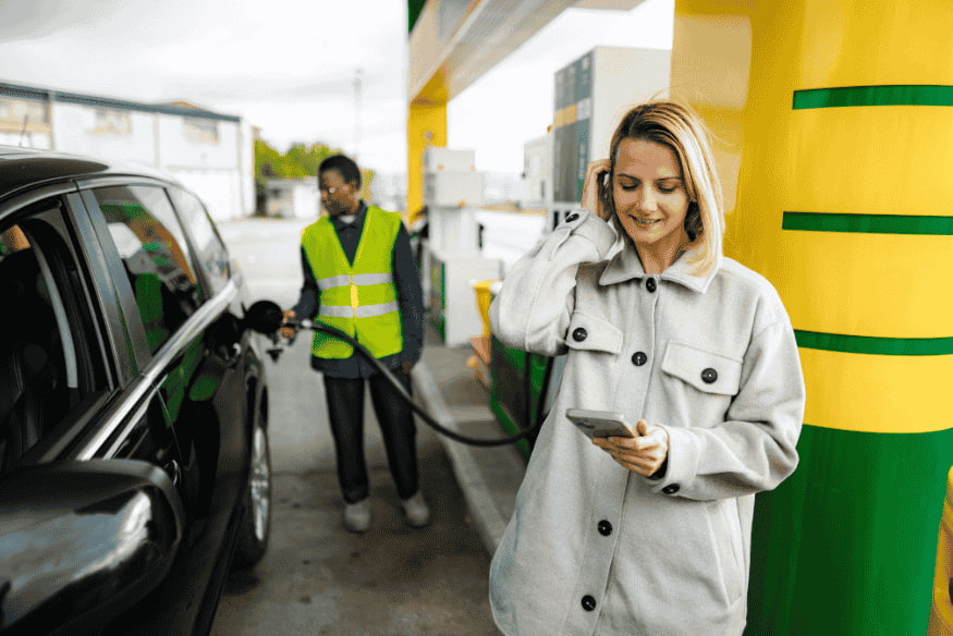 Frau an der Tankstelle mit Smartphone neben Auto beim Tanken als Beispiel für variable Kosten im Alltag und steigende Mobilitätsausgaben